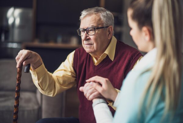 Elderly man with glasses sitting with nurse, holding a walking stick